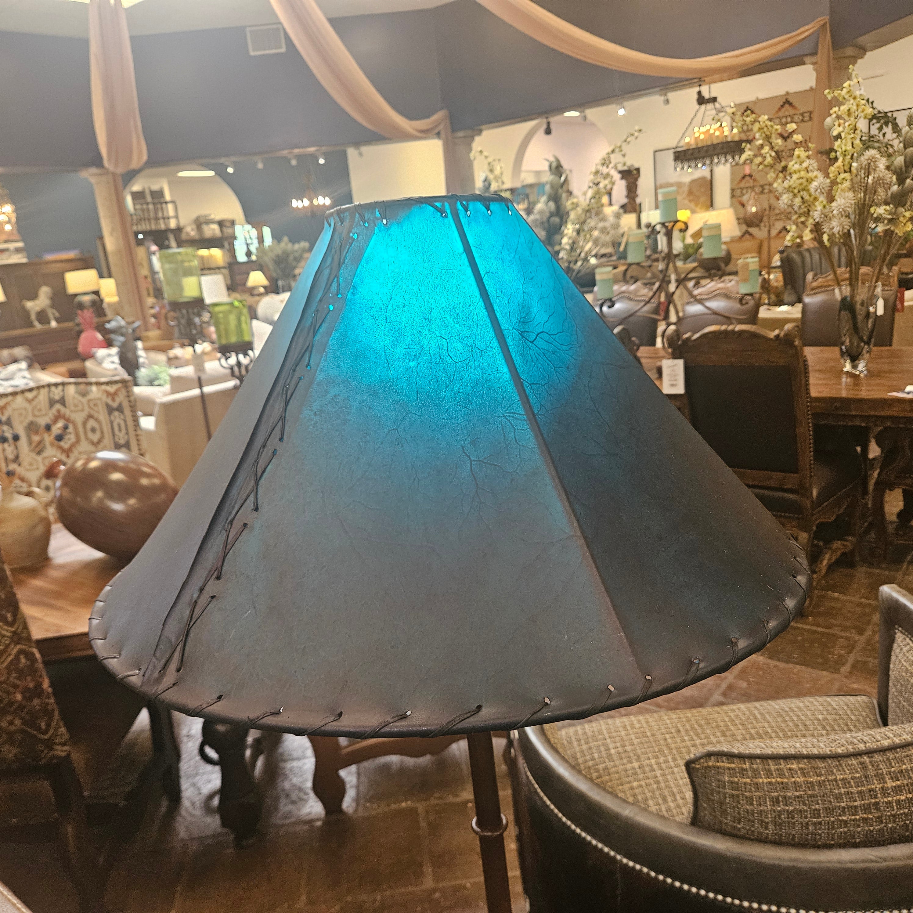 A large conical lampshade with a dark charcoal gray leather-like finish and visible whipstitching along the edges, shown lit with a bright blue glow in a furniture store interior.