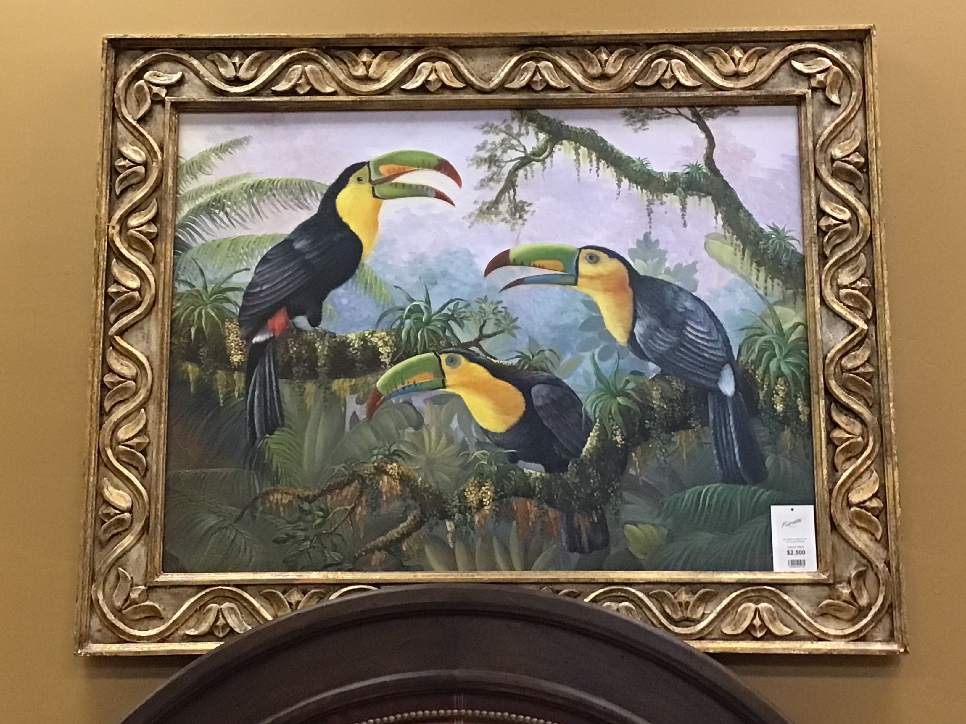 Framed painting of toucans in a jungle setting on a beige wall.