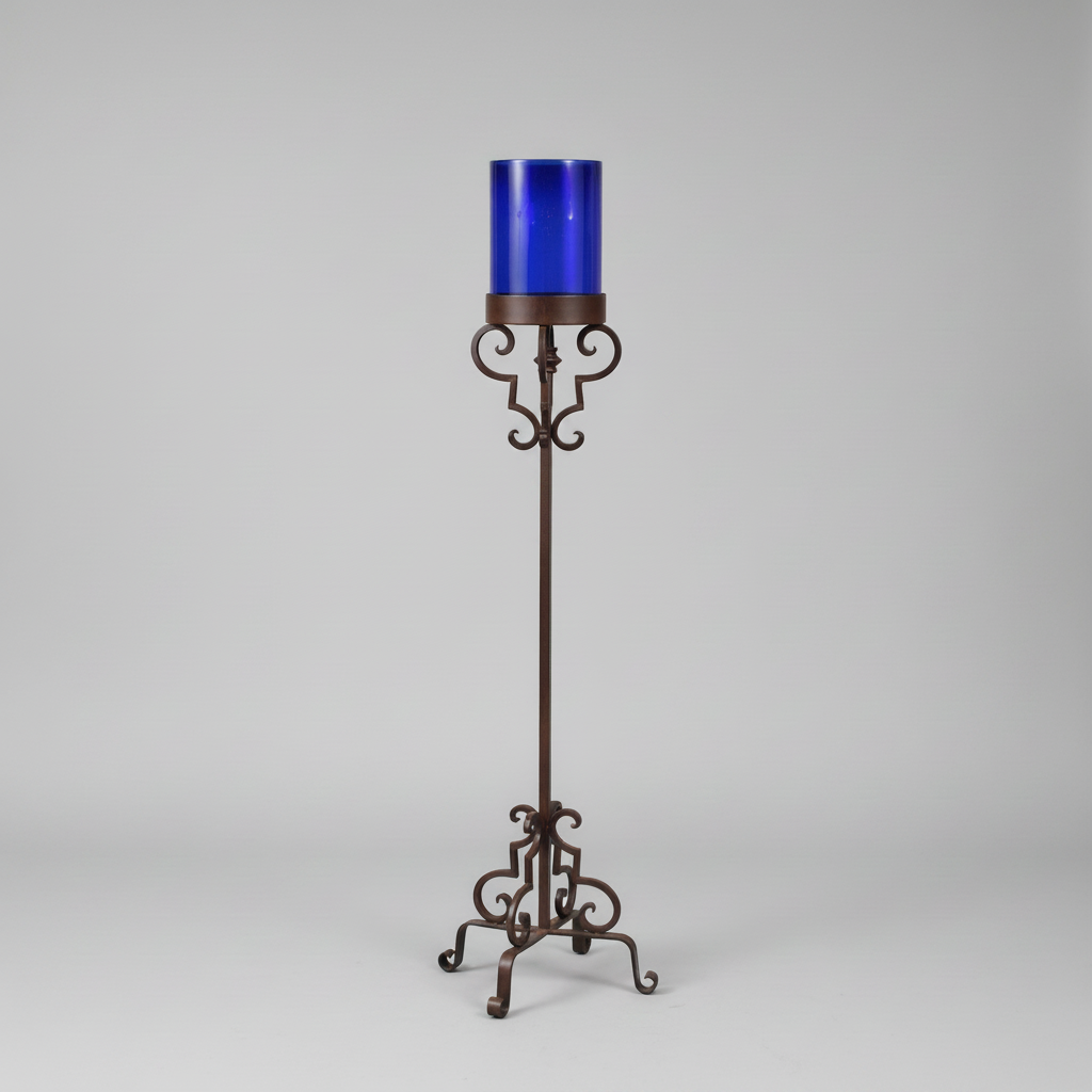 A tall decorative metal candle holder with an ornate dark brown wrought-iron style stand and a translucent cobalt blue glass cup, shown on a light gray studio background.