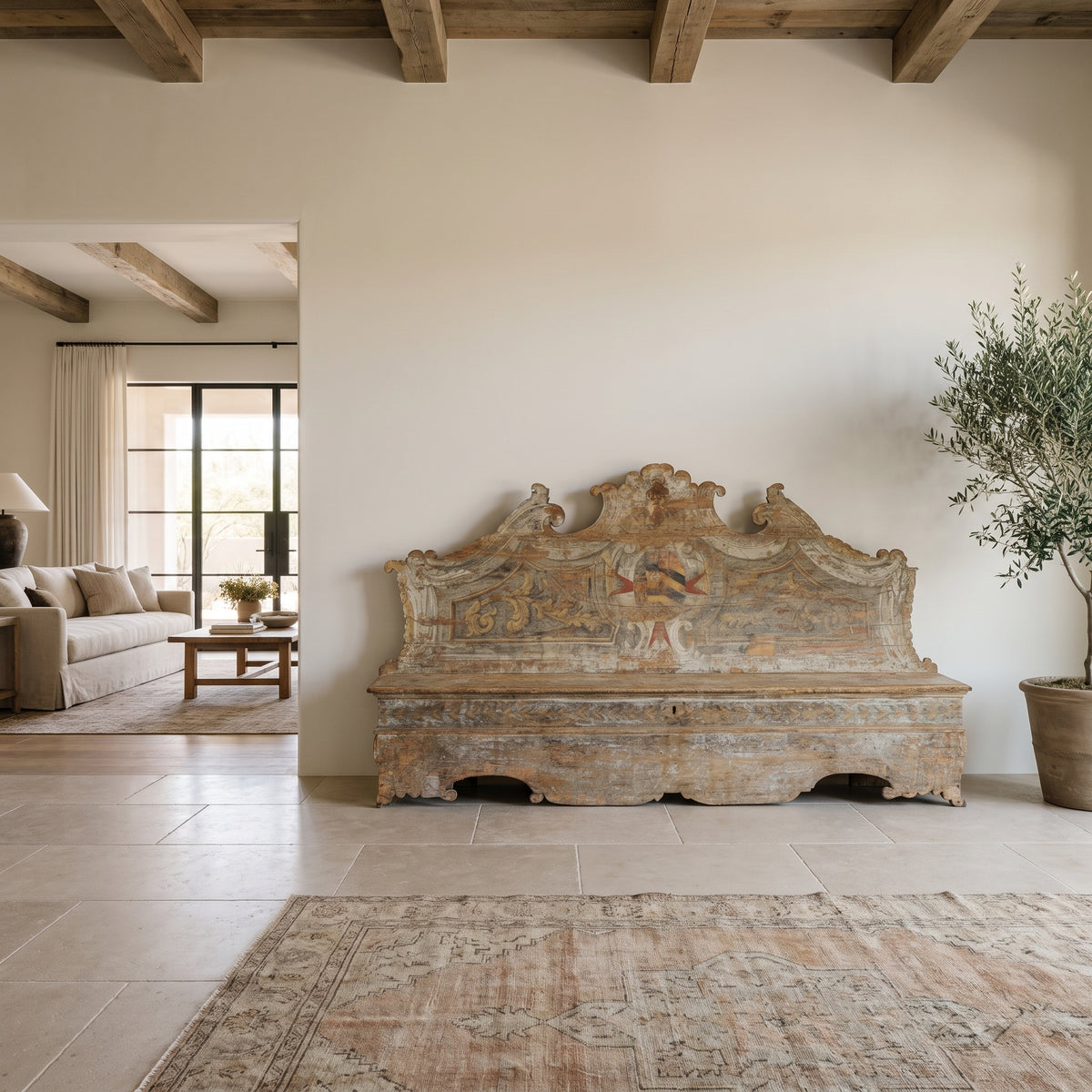 18th-century Italian painted trunk bench with a carved, scrolled crest and heraldic cartouche, displayed in a modern Scottsdale entryway with limestone floors, wood ceiling beams, a vintage rug and an olive tree.