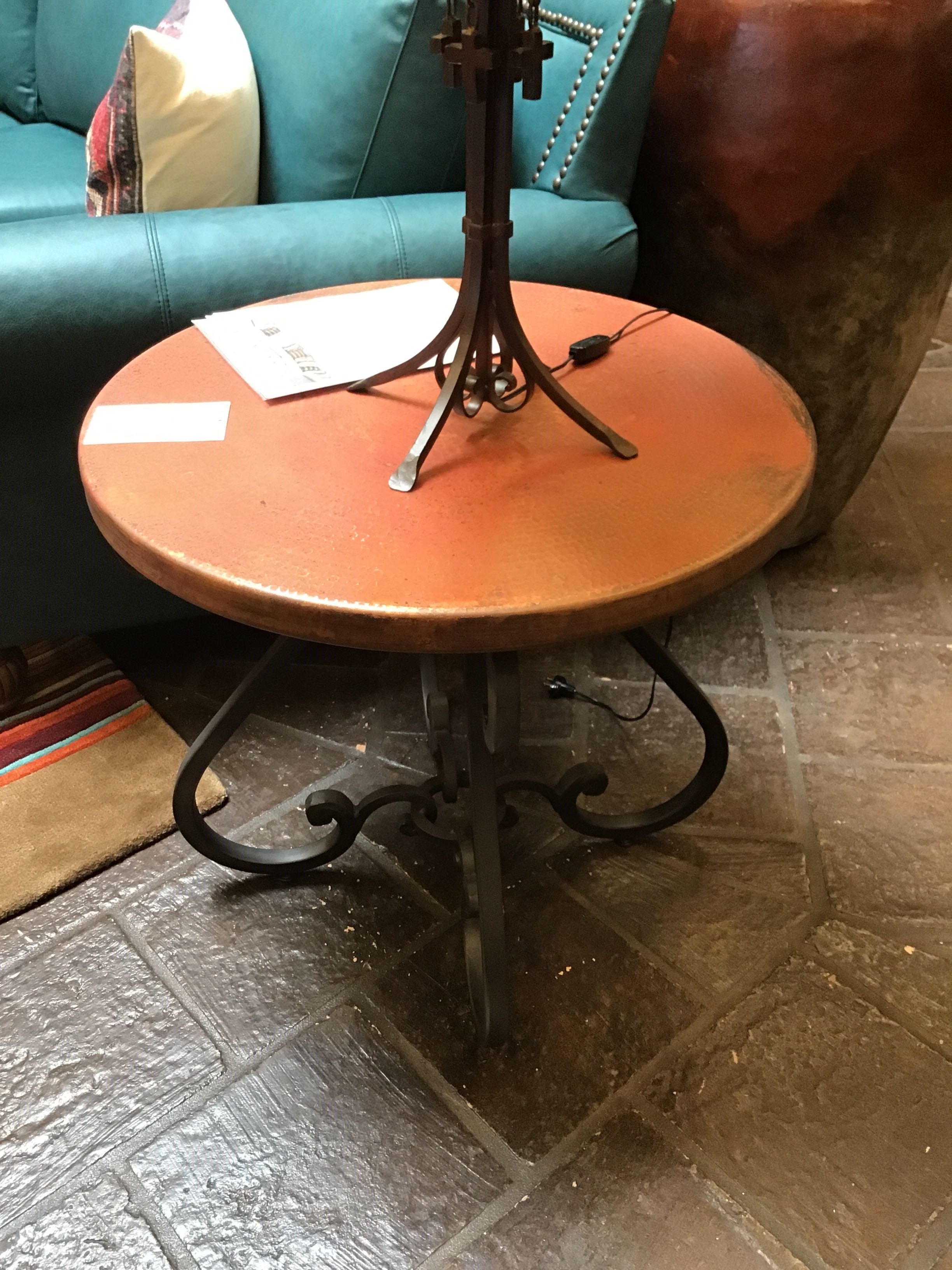 Round wooden table with metal legs on a stone floor next to a teal sofa.