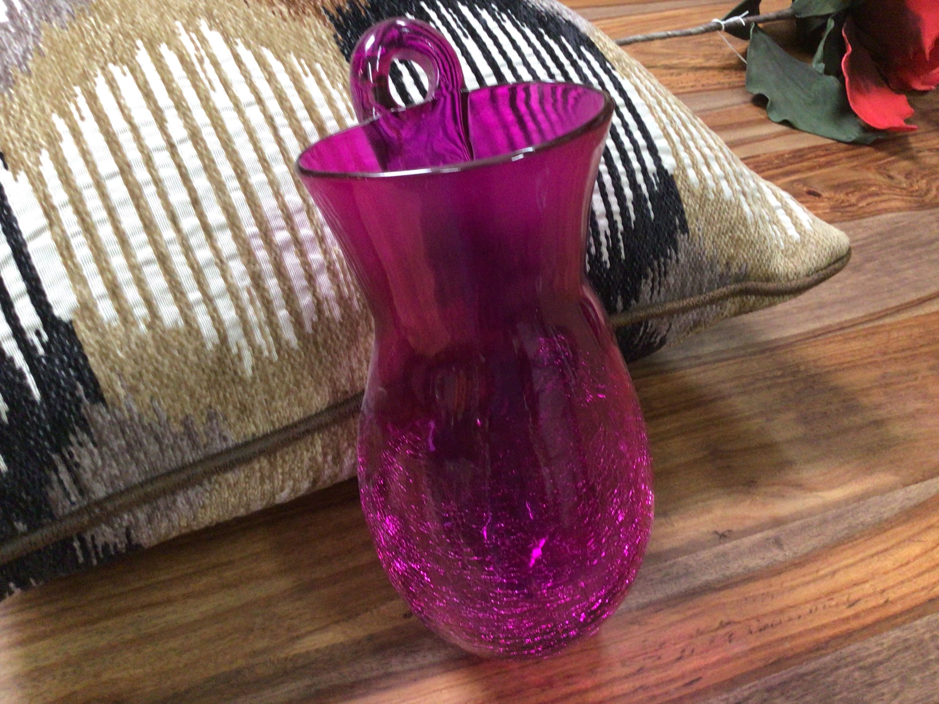 Purple glass vase on a wooden surface with a patterned cushion in the background