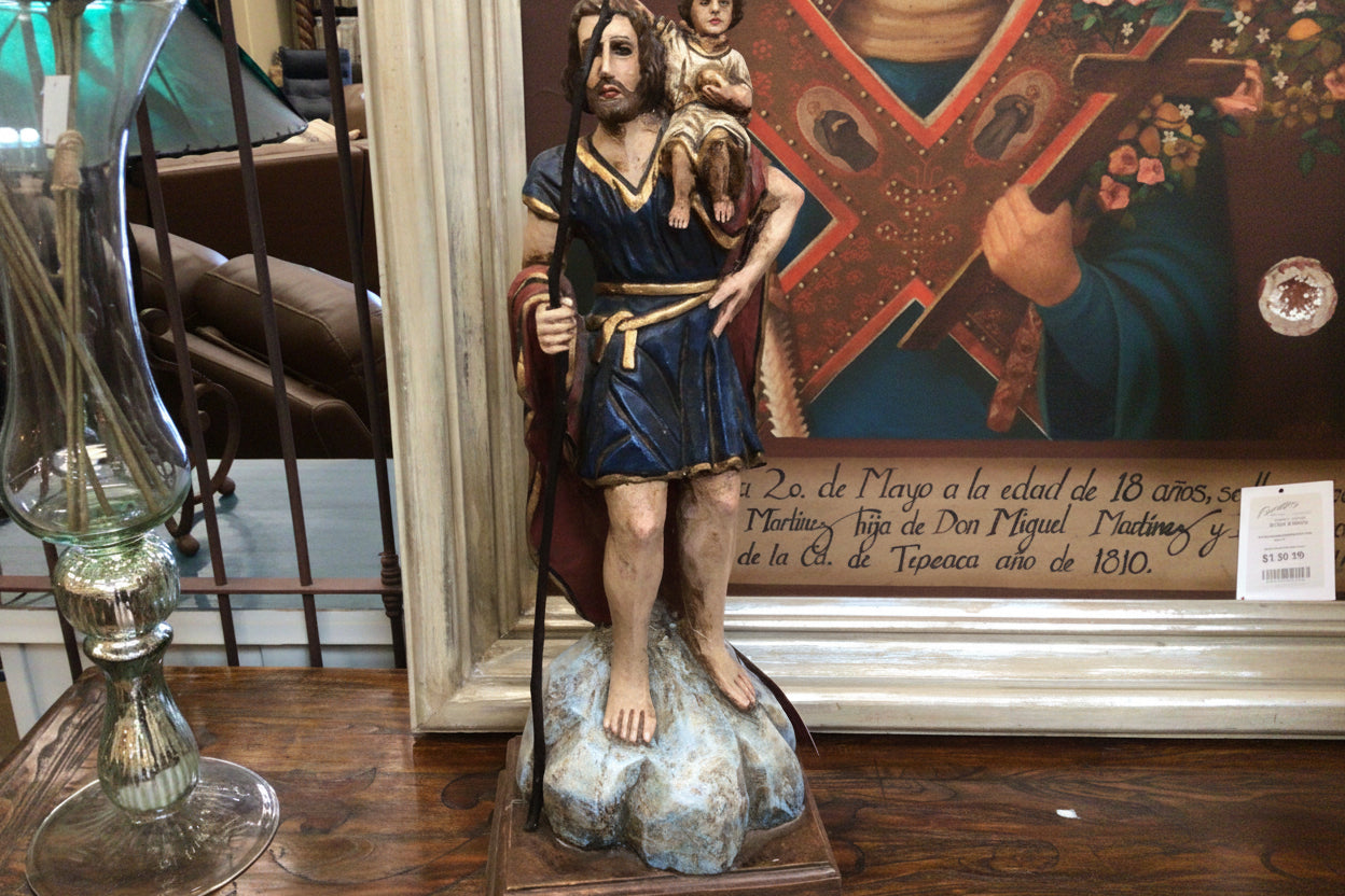Statue of a religious figure holding a cross on a wooden table with a painting in the background.