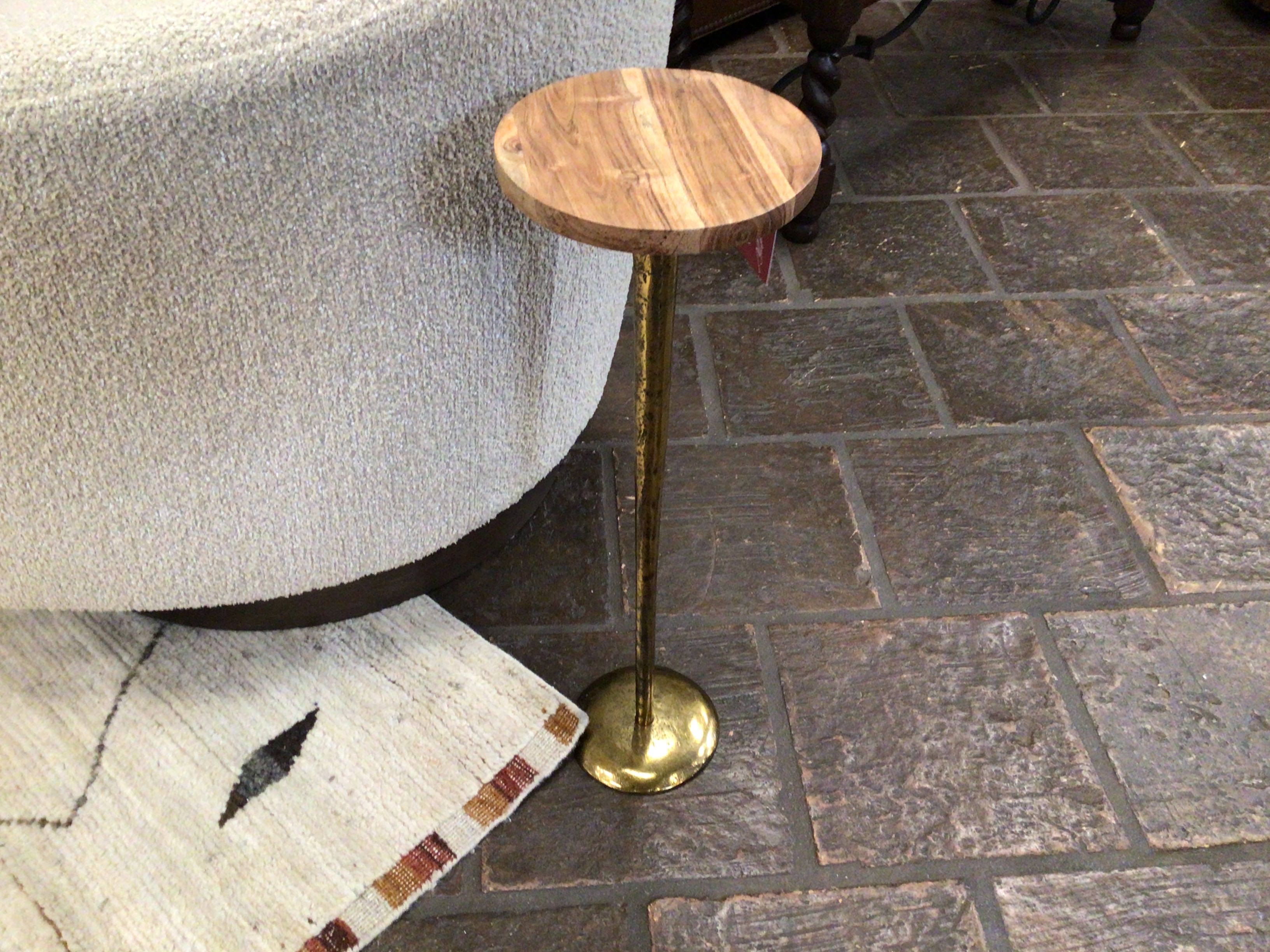 Small round wooden table with brass base on a stone floor next to a sofa.