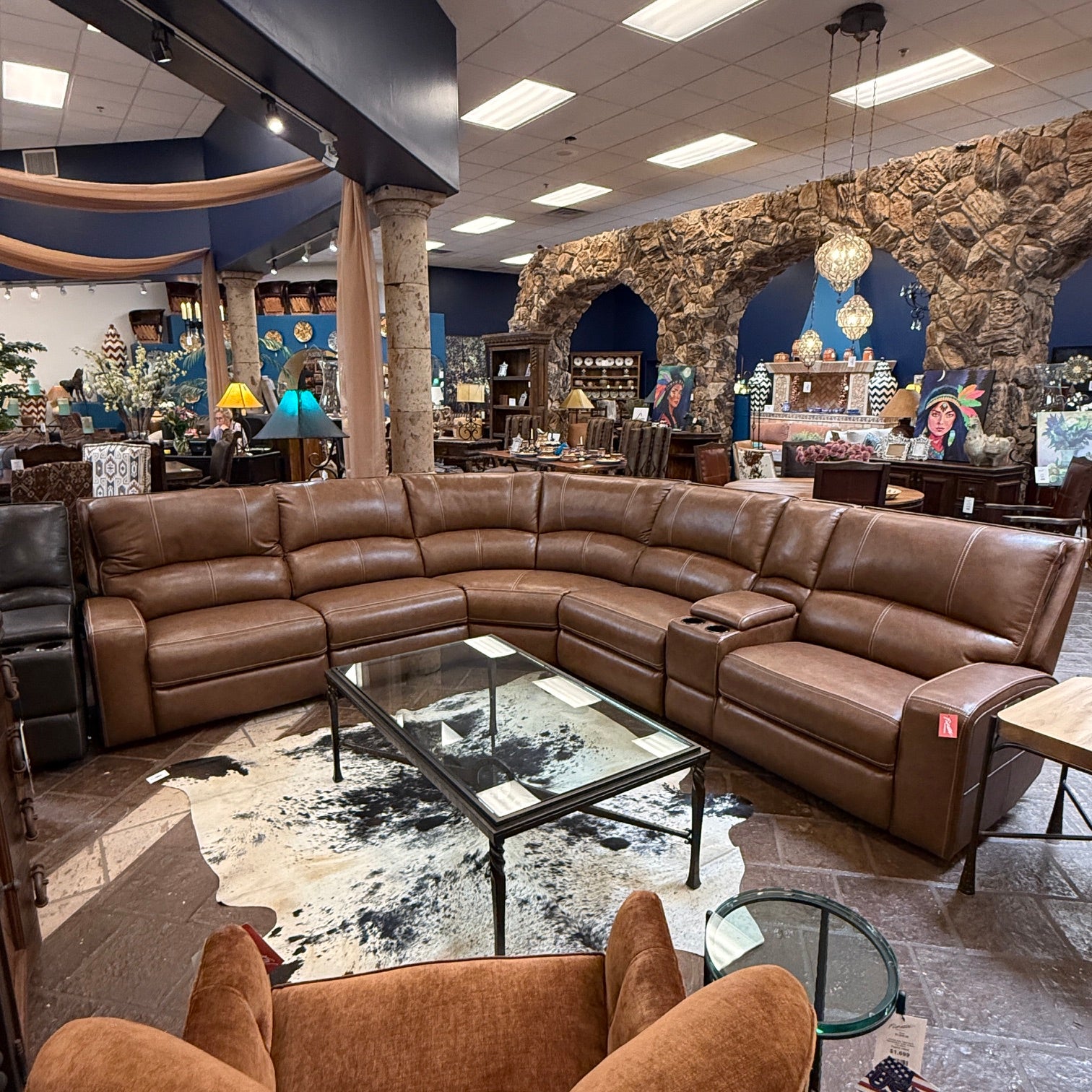A large brown leather sectional sofa arranged in a U-shape in a furniture showroom, with a glass-top coffee table and patterned rug in front.