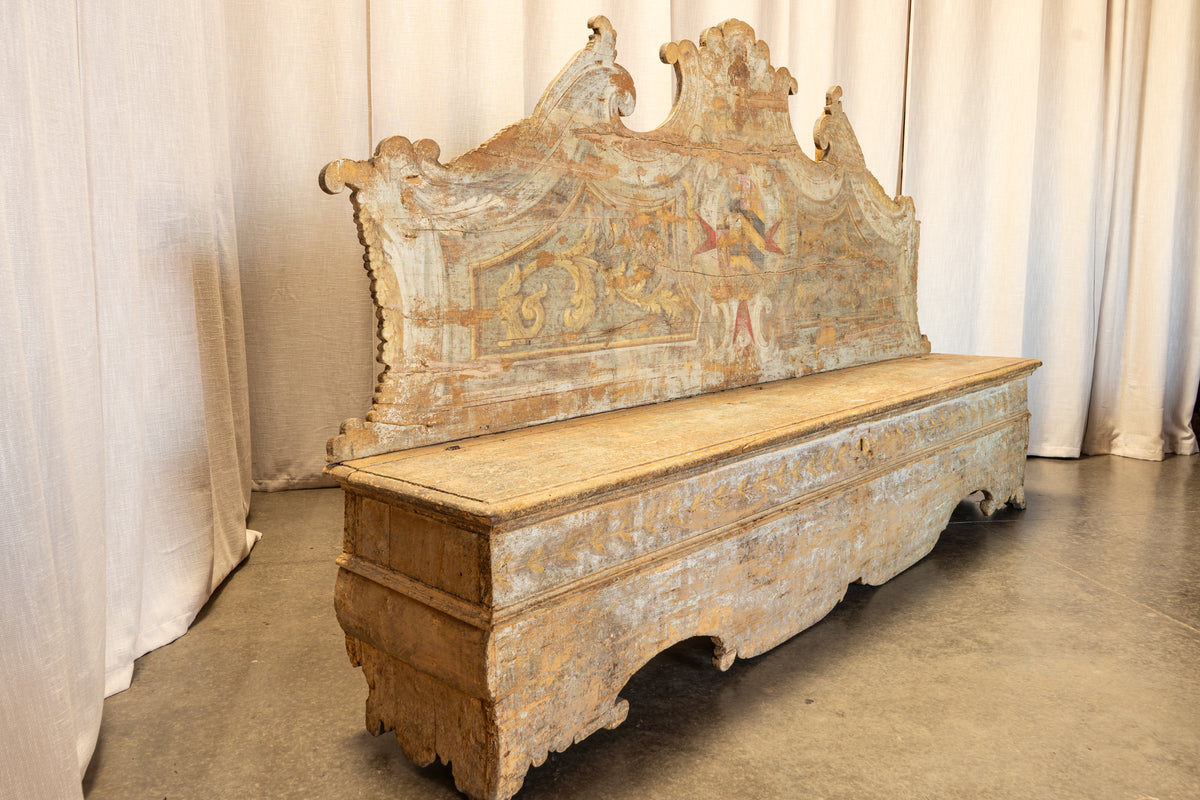 An antique wooden chest or bench with a tall, ornate painted backrest, photographed indoors against draped white fabric and a concrete floor.