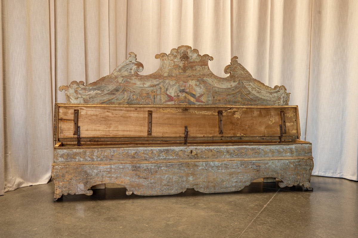 An antique wooden chest or bed-like storage piece with a highly decorative carved crest and faded blue-gray painted lower section, photographed indoors against draped cream curtains.