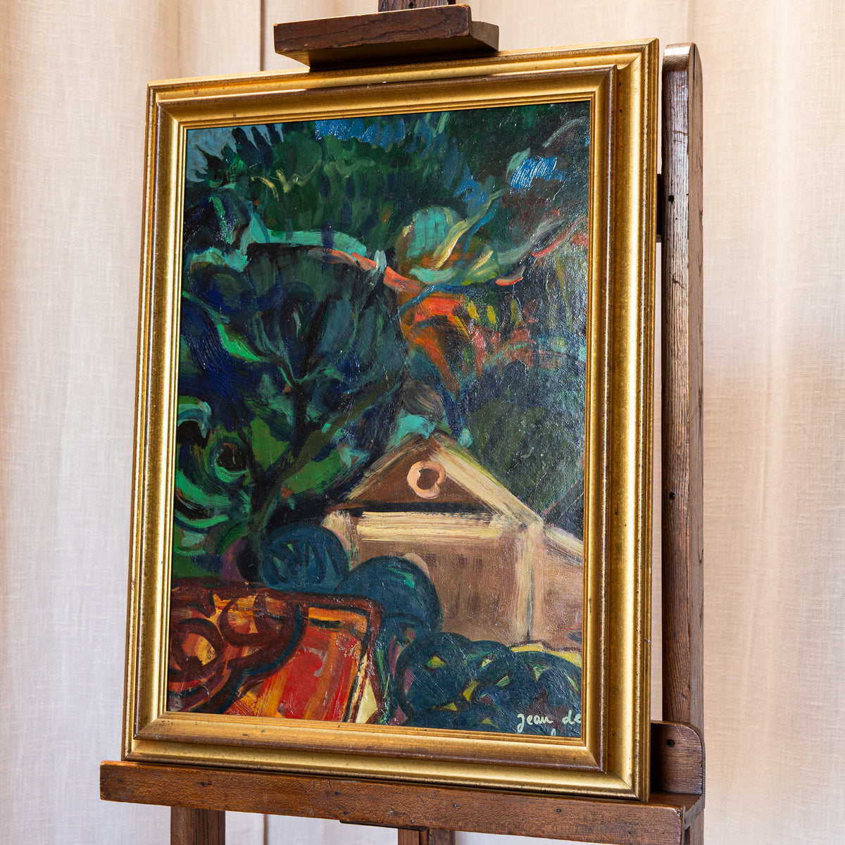 A framed painting depicting an abstract outdoor scene with dark green foliage, blue shapes, and a beige house, displayed on a wooden easel against a light beige curtain background.