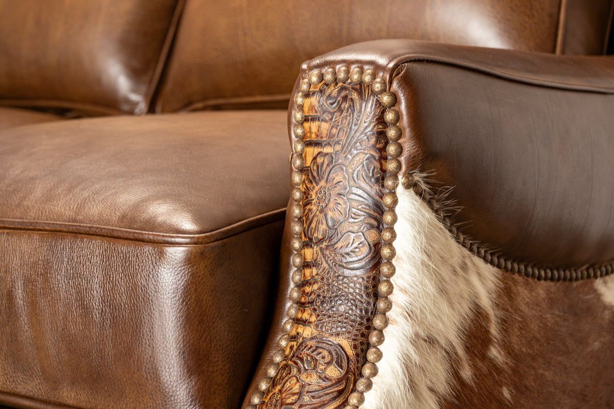 Close-up of a brown leather sofa armrest with carved floral tooling, a border of metallic nailheads, and a brown-and-white cowhide panel.