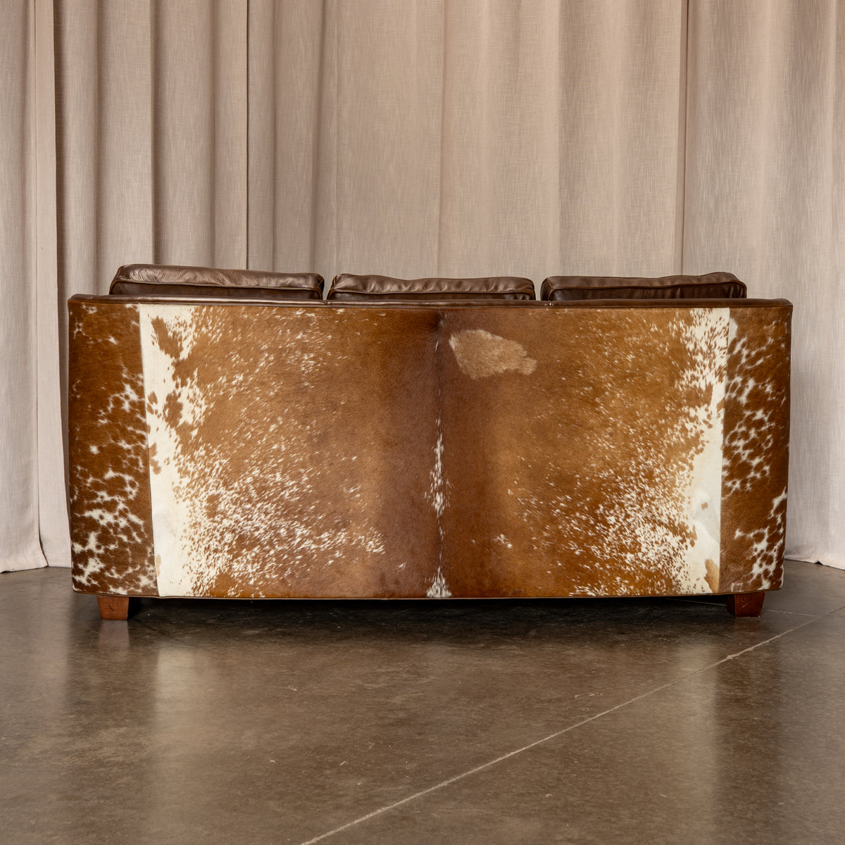A three-seat sofa upholstered in brown and white cowhide, positioned against a neutral beige curtain backdrop on a polished concrete floor.