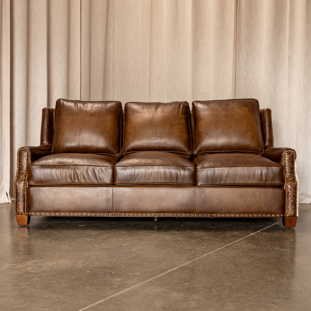 A brown leather three-seat sofa with rolled arms, nailhead trim, and wooden legs, positioned against a beige curtain backdrop on a concrete floor.