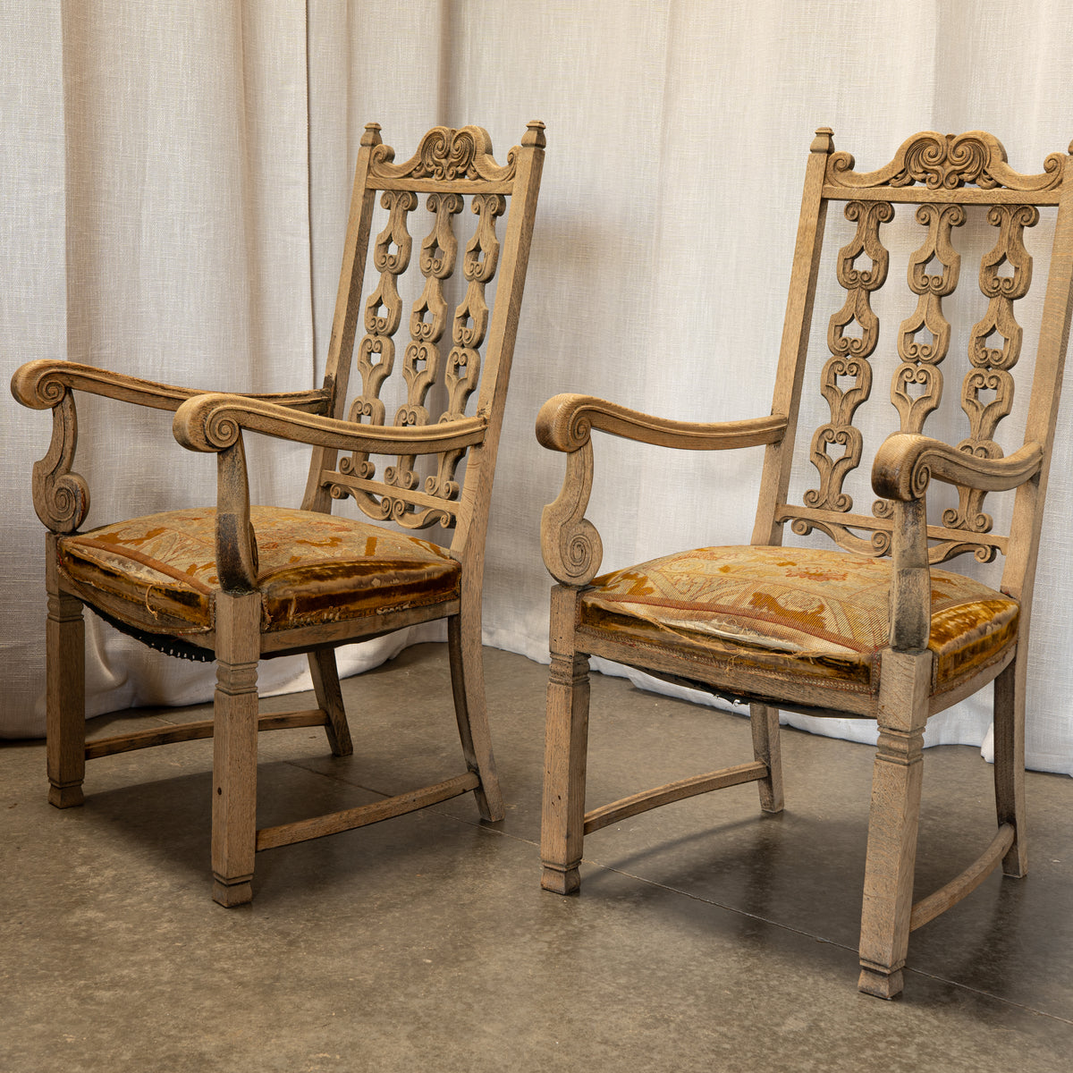 Two ornate carved wooden armchairs with high pierced backs and patterned upholstered seats, shown against a draped fabric backdrop and concrete floor.