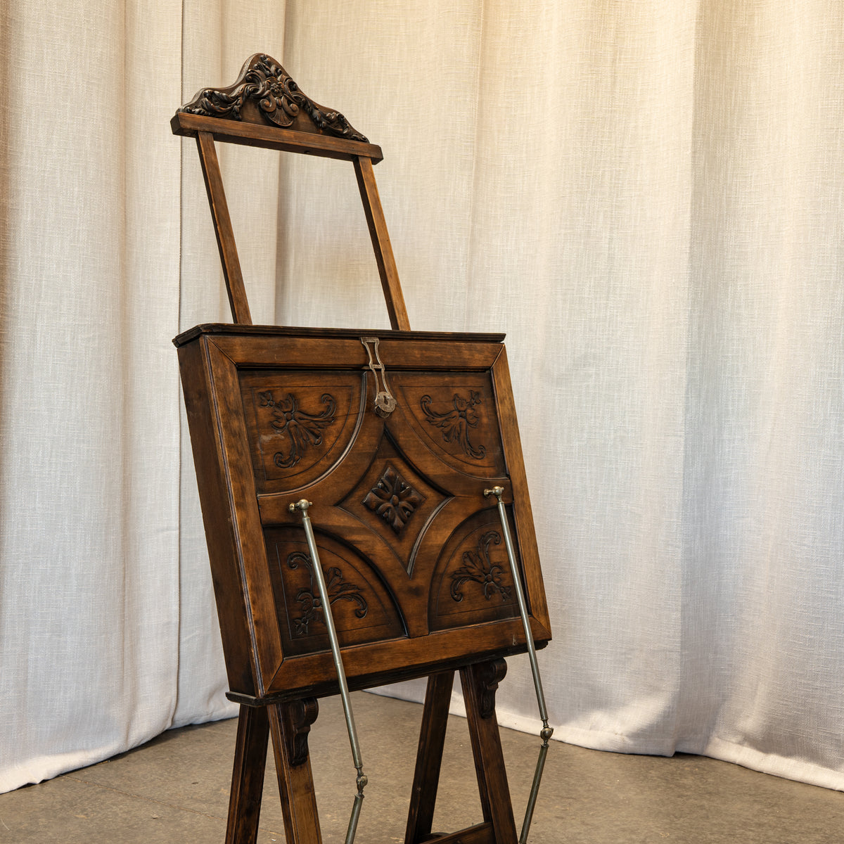 A dark brown wooden easel with ornate carved details on the front panel and a decorative top frame, shown standing against a draped off-white fabric backdrop.