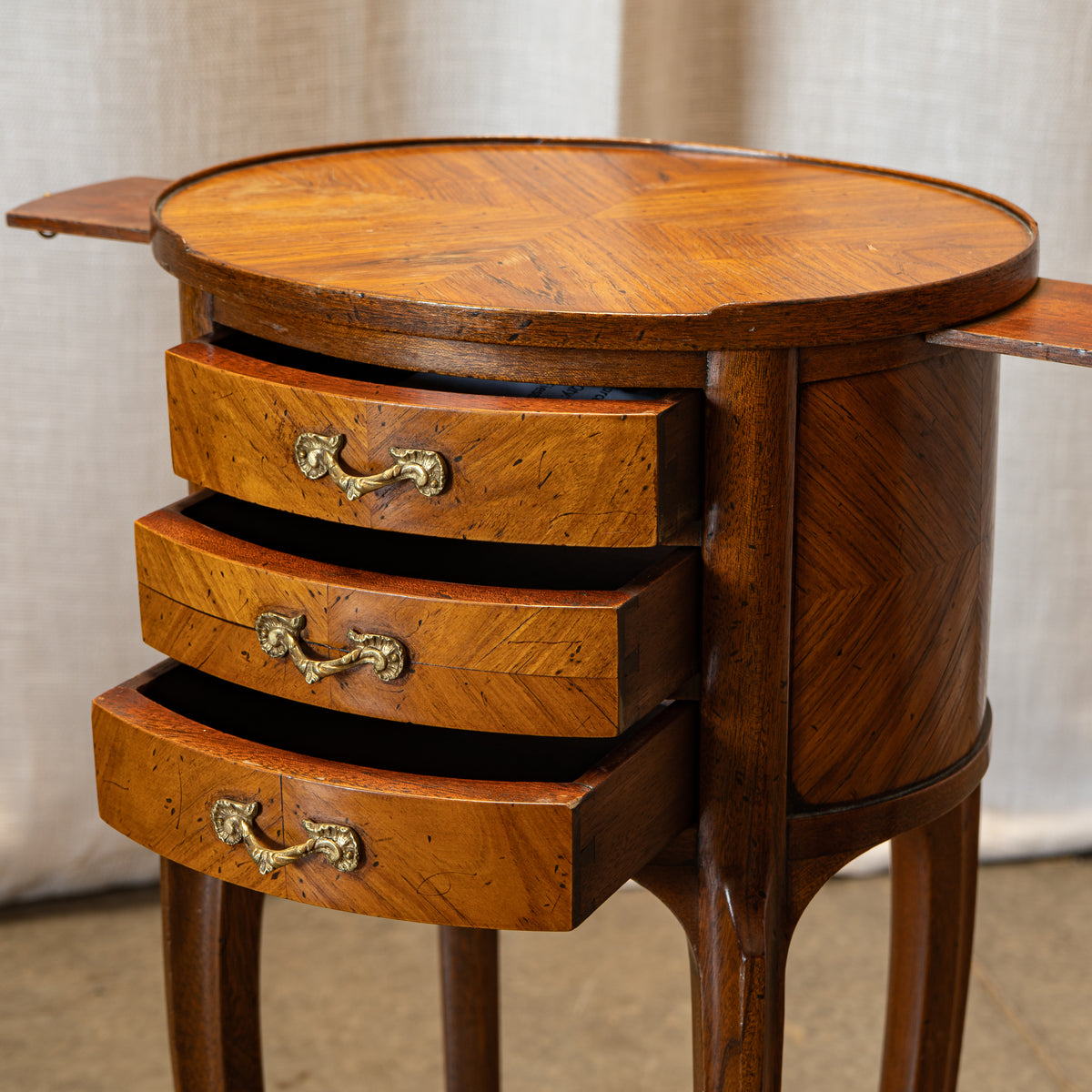 An antique wooden side table with three drawers, featuring a round top, herringbone wood grain, and brass handles, set against a neutral draped fabric background.