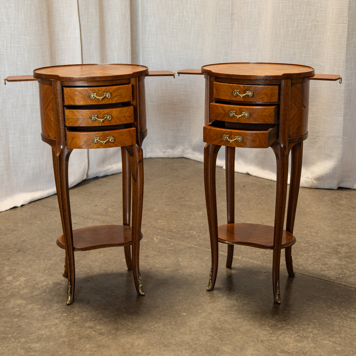A pair of vintage wooden nightstands with three drawers, brass handles, curved legs, and a lower shelf, set against a draped fabric backdrop.