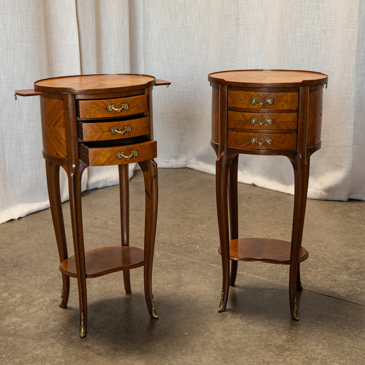 A pair of vintage wooden nightstands with three drawers, brass handles, and curved legs, set against a neutral draped fabric backdrop.