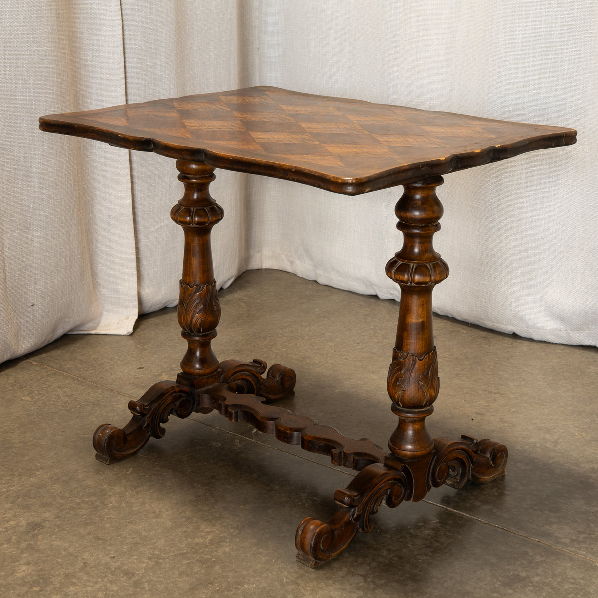 An antique dark brown wooden table with a checkered inset top, carved turned legs, and a decorative stretcher base, set against a neutral draped fabric backdrop.