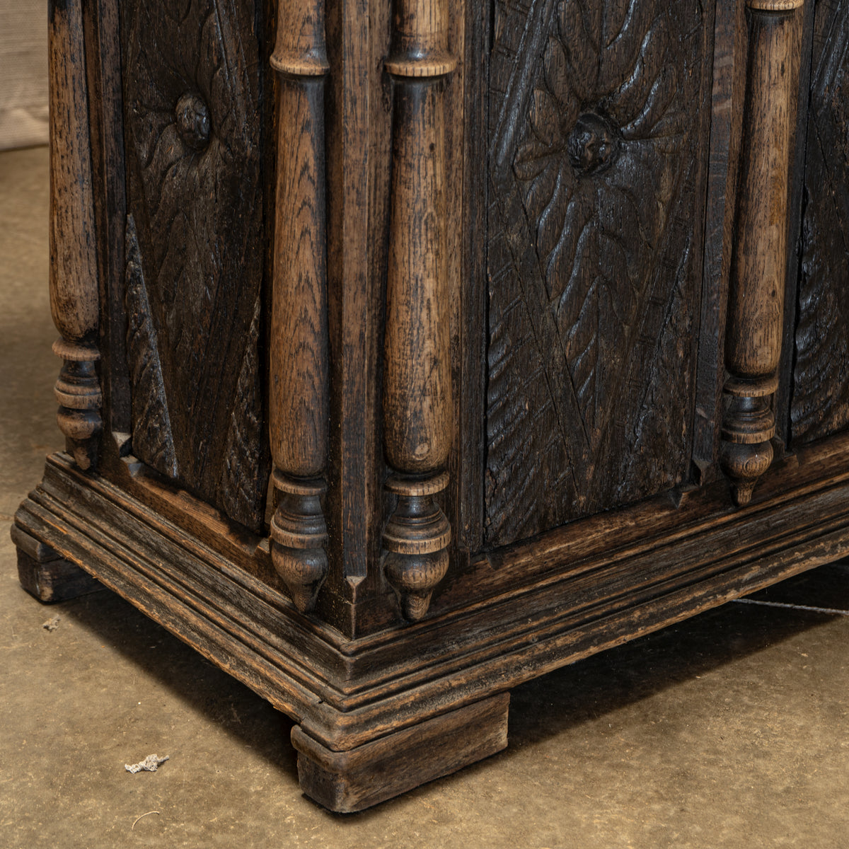 Close-up of a dark carved wooden cabinet corner with turned posts and a distressed finish, shown on a concrete floor.