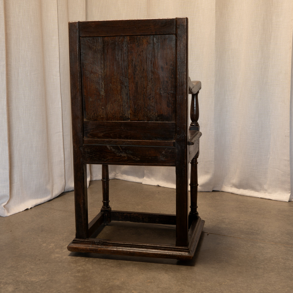A dark brown wooden armchair with a high paneled backrest and turned legs, positioned against a backdrop of draped off-white fabric curtains.