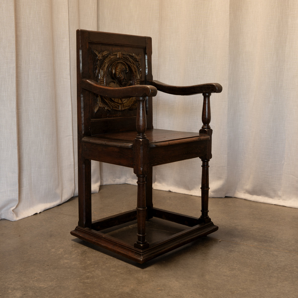 A dark brown carved wooden armchair with a square frame and decorative backrest, set against a neutral draped fabric backdrop.