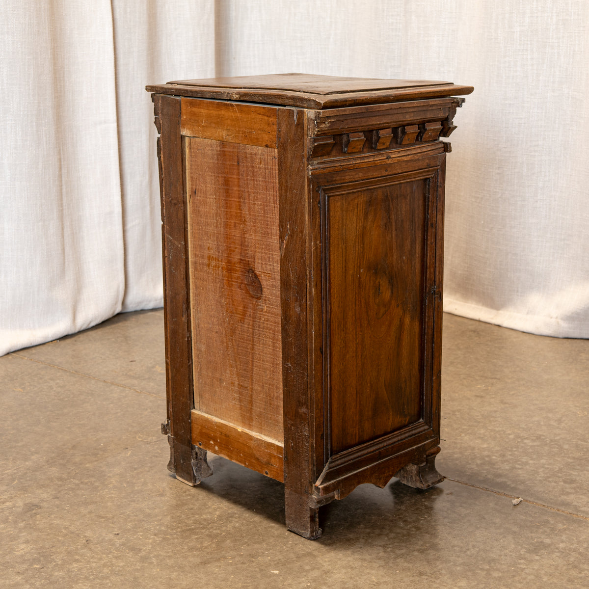 An antique wooden side table or cabinet with carved details and a hinged top, shown against a neutral draped fabric backdrop.