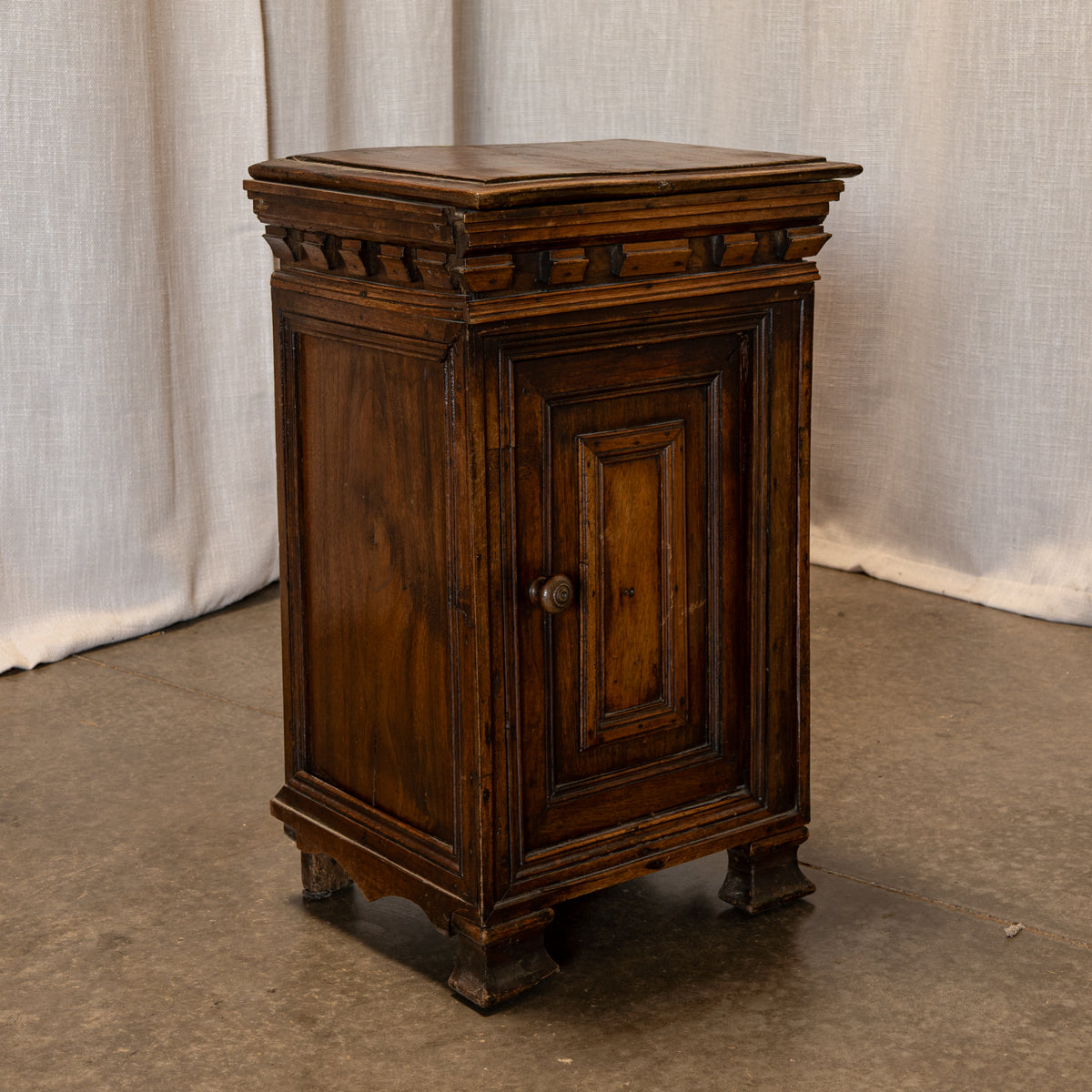 An antique dark brown wooden cabinet with a single paneled door, decorative trim, and small feet, set against a draped fabric backdrop.
