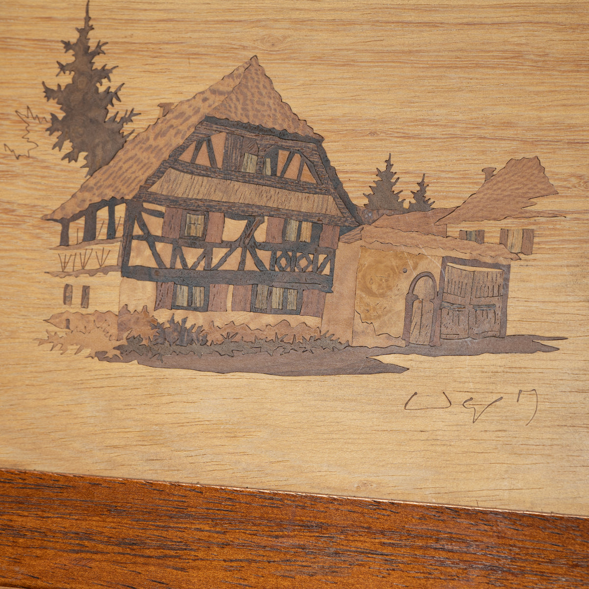 A close-up of a wooden surface featuring an engraved or burned depiction of a traditional European half-timber house with trees and a smaller building in the background. The wood grain is visible, with a darker brown band along the bottom edge.