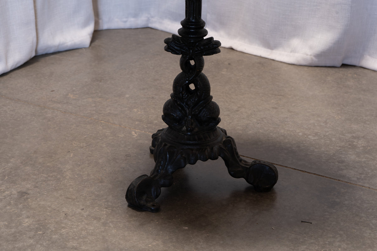 Close-up of an ornate black cast iron table base with three legs, set on a concrete floor with draped white fabric in the background.
