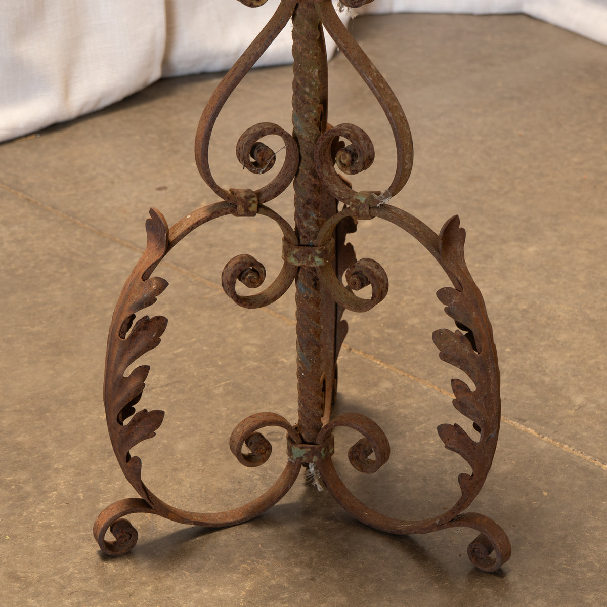 Rustic wrought iron stand with ornate scrolled details and leaf-like flourishes, photographed on a stone floor with a white fabric surface in the background.