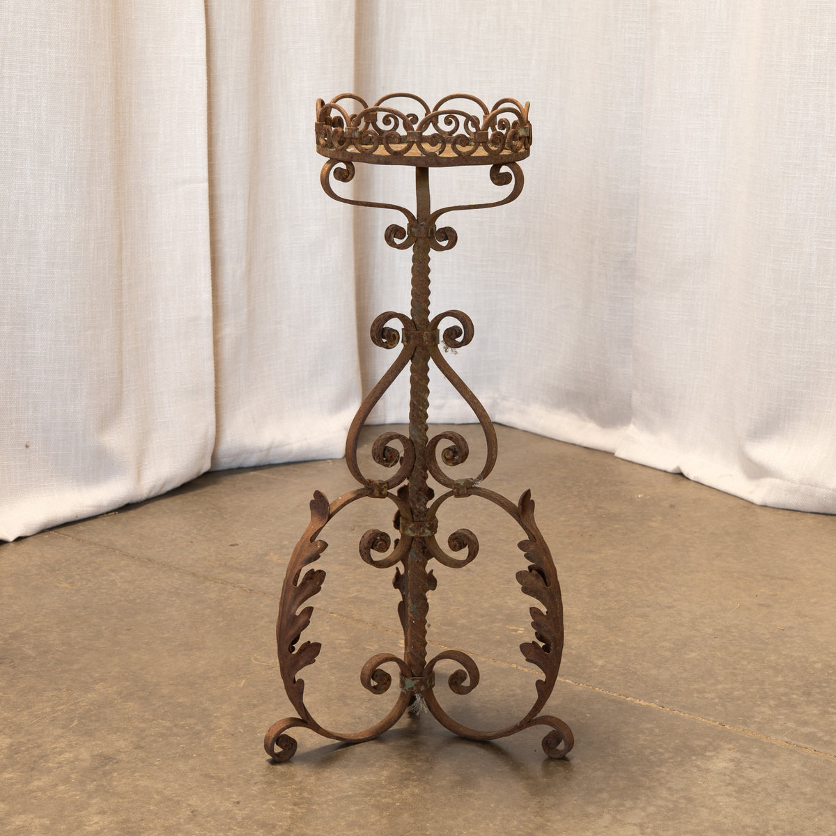 An ornate rusted metal candle holder stand with a decorative top and scrollwork base, photographed against draped white fabric and a concrete floor.