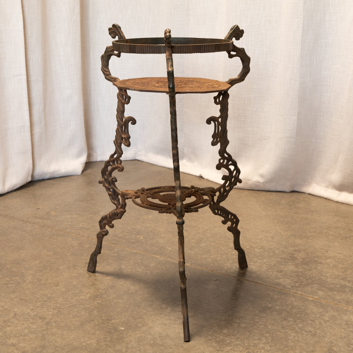 An ornate vintage cast iron plant stand with three tiers, featuring scrolled legs and a dark rusted finish, set against a draped white fabric backdrop.