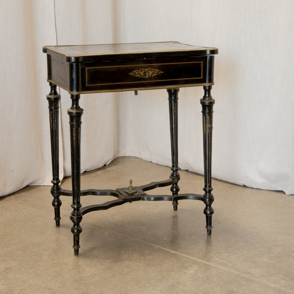 An antique black wooden writing desk with gold-toned trim, turned legs, and a central brass drawer pull, set against a draped white fabric backdrop.