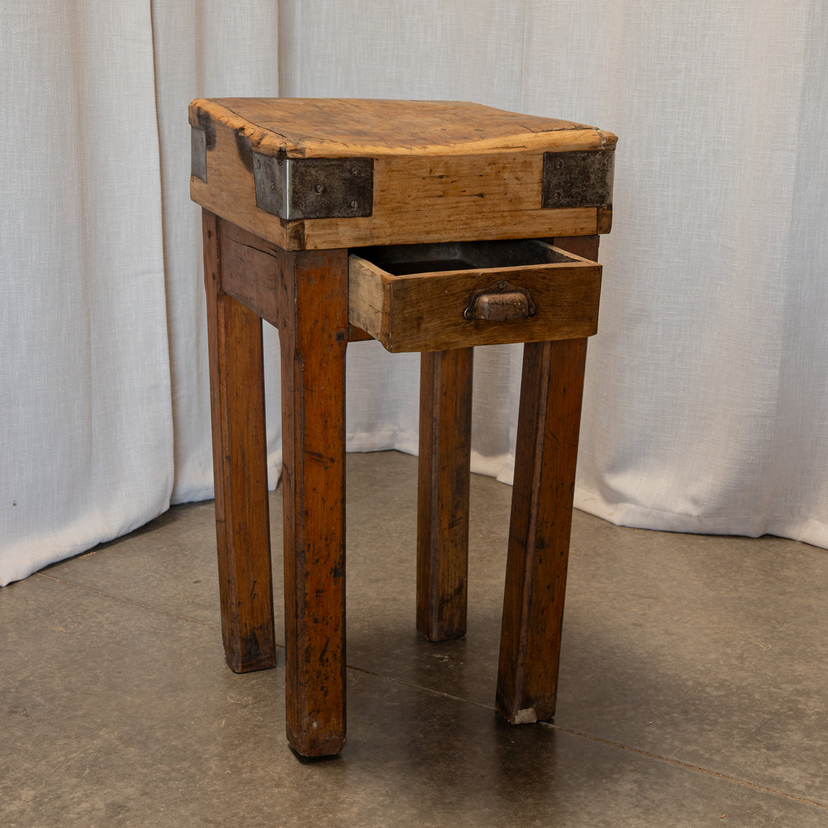 A rustic wooden chest-topped stool with a single pulled-out drawer, photographed against a draped white fabric backdrop and concrete floor.