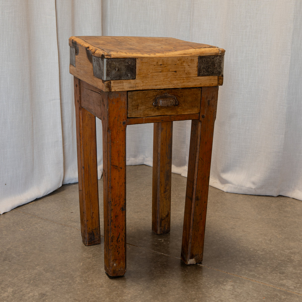 A rustic wooden stool with a square top, single drawer, and four legs, featuring dark metal corner brackets and a small metal pull handle, set against a draped white fabric backdrop.