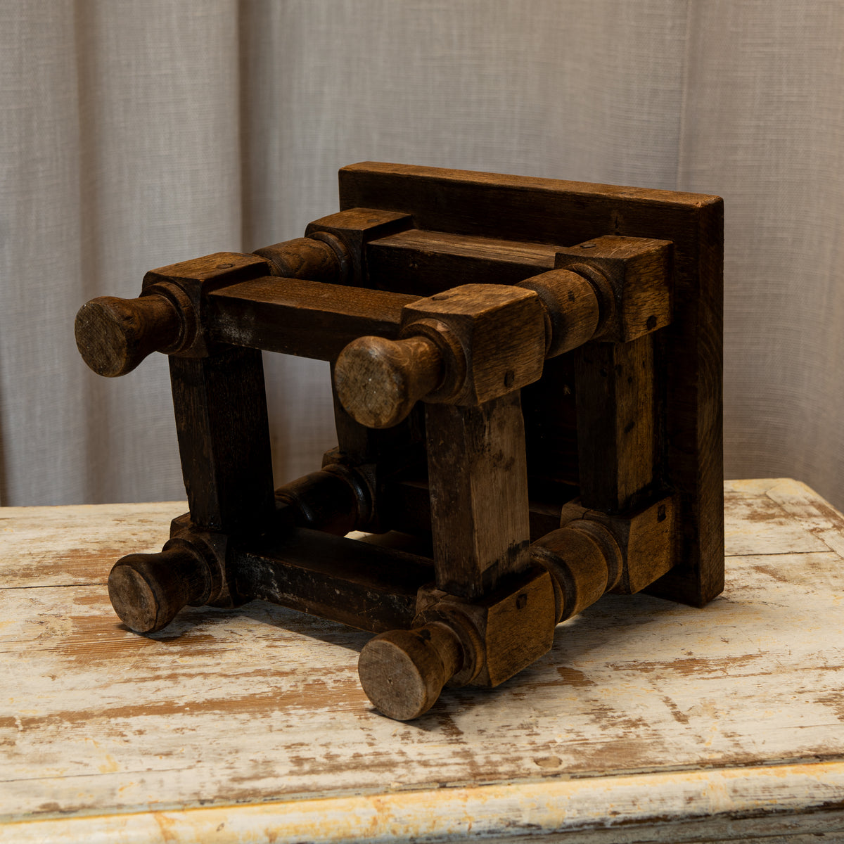 A rustic dark brown wooden stool with round turned legs and a thick backrest, shown on a distressed white wooden table against a neutral curtain background.