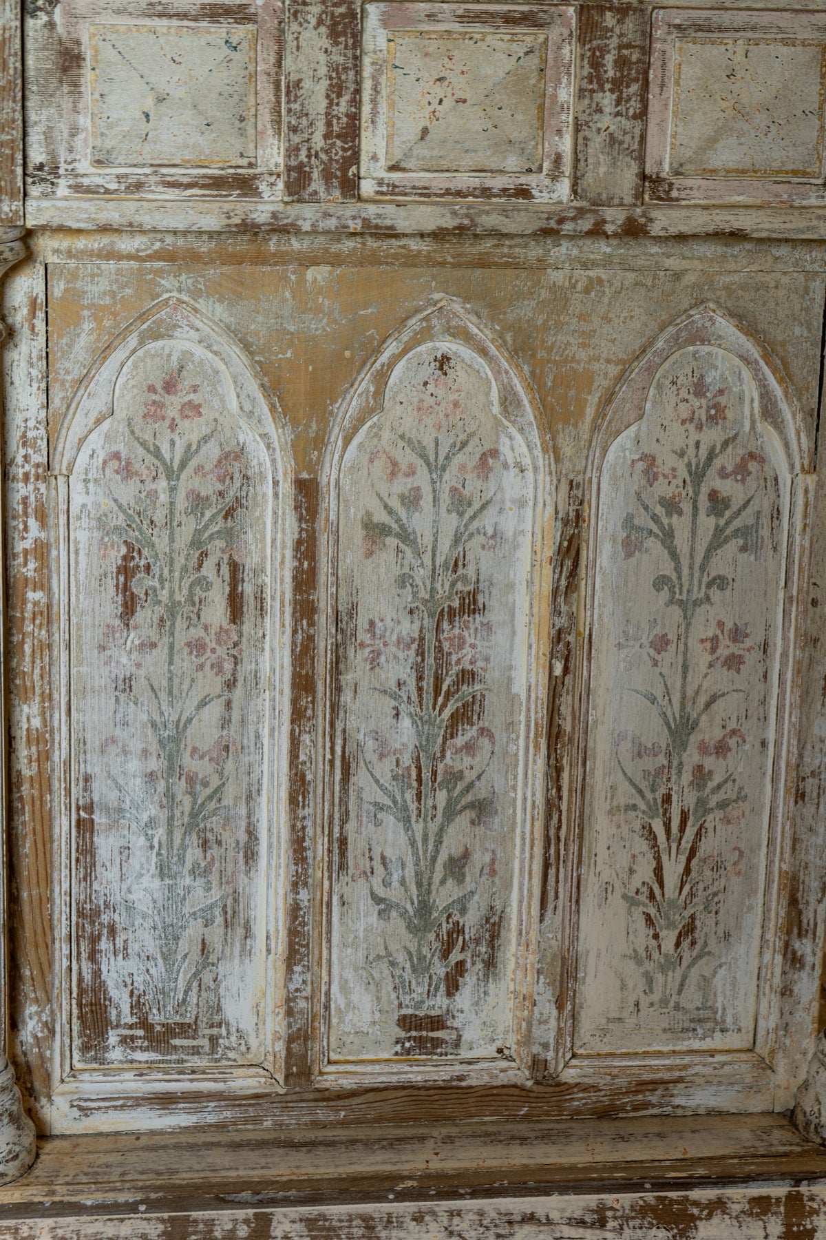 An antique painted wooden panel with three arched floral motifs in muted pink, green, and gray tones over a heavily distressed beige and brown surface.