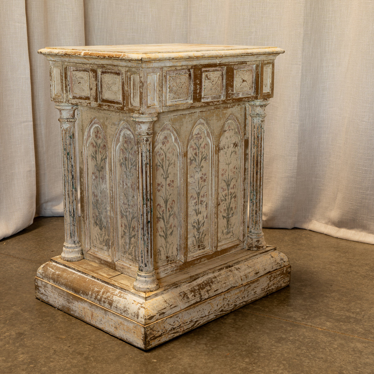 An antique-style distressed wooden altar or lectern with a weathered beige and brown finish, featuring carved panel details and fluted columns, set against a neutral draped fabric backdrop.