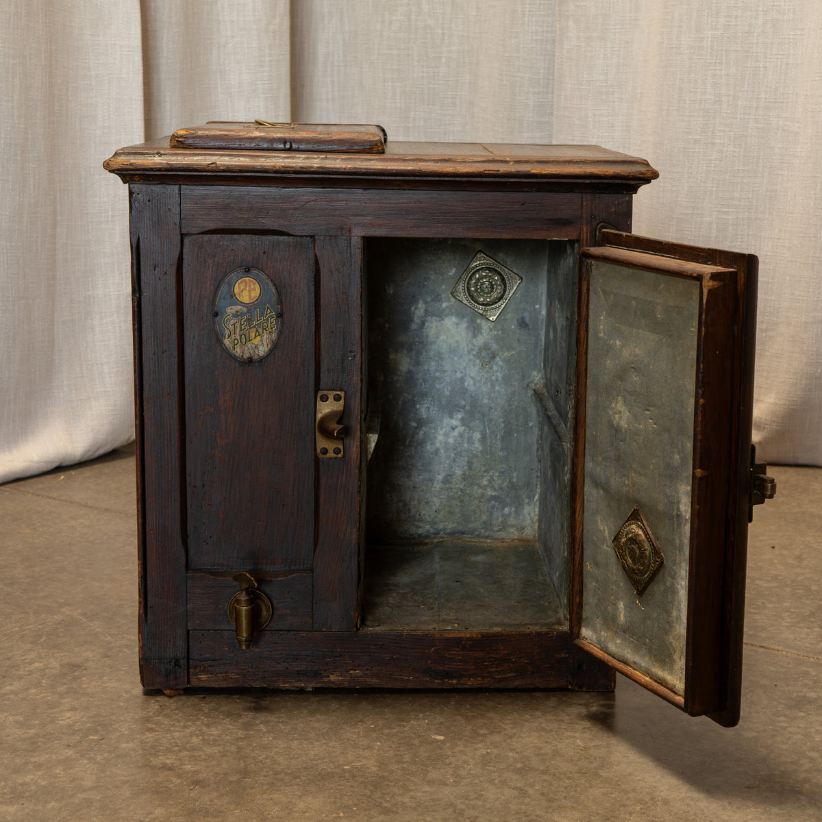An antique dark wood chest or cabinet with one door open, showing a metal-lined interior. The cabinet has brass hardware, a small round decorative emblem on the door, and a latch on the front.