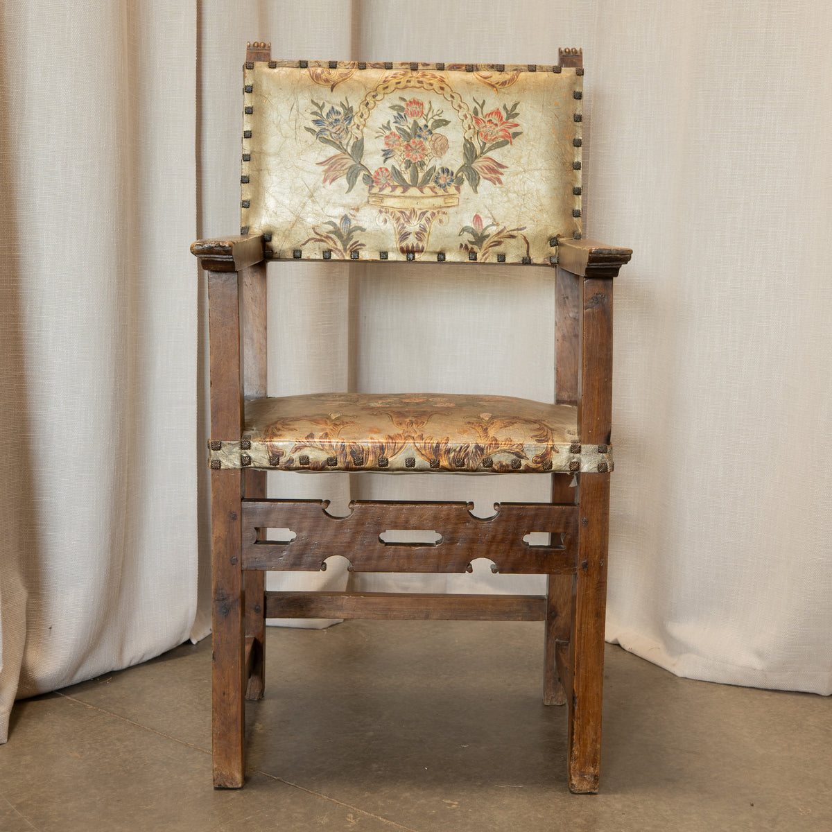 A vintage wooden armchair with a floral tapestry upholstered backrest and seat cushion, featuring brass nailhead trim, set against a neutral draped fabric backdrop.