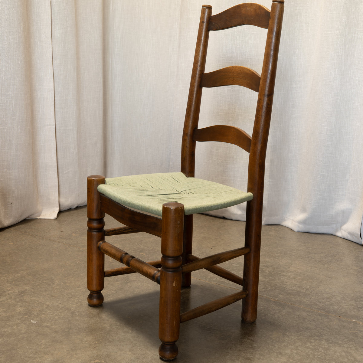 A wooden ladder-back dining chair with a woven light green seat, positioned against a neutral backdrop of draped fabric curtains and a concrete floor.
