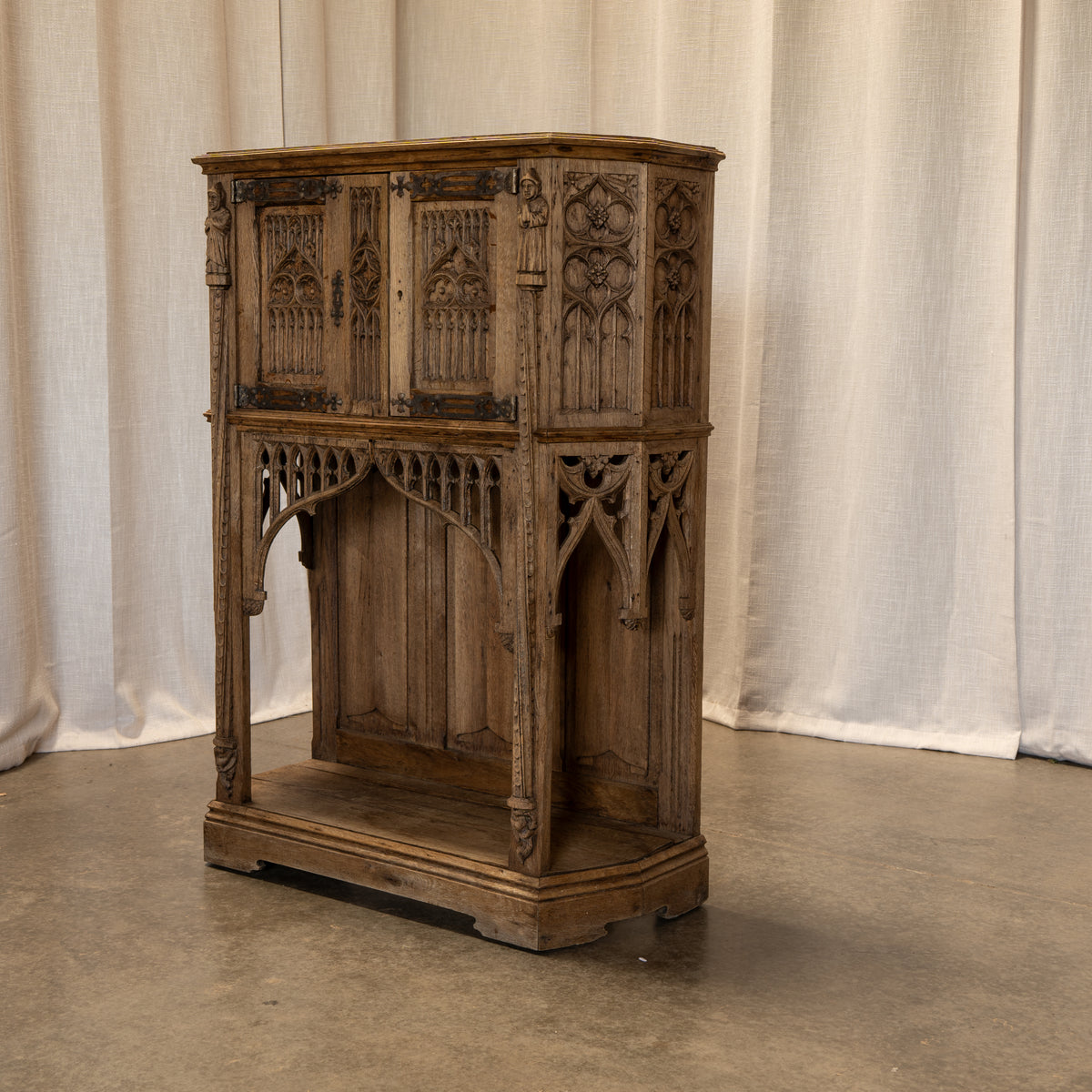 An ornate antique wooden cabinet with Gothic architectural details, including carved panels, arched supports, and a lower shelf, photographed against a draped off-white fabric backdrop on a concrete floor.
