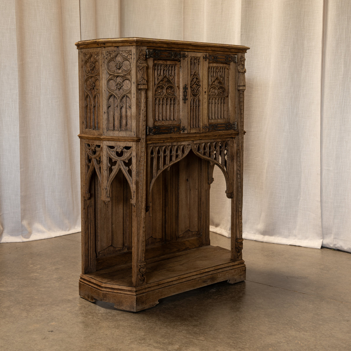 An ornate Victorian Gothic-style wooden cabinet with carved details, dark weathered brown finish, and brass hardware, set against a draped off-white fabric backdrop.