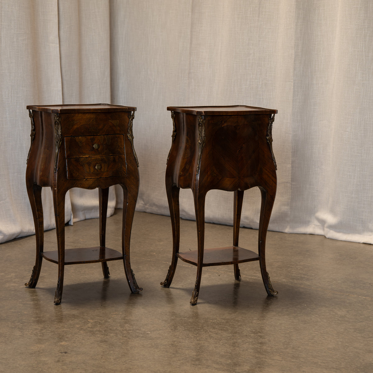 A pair of antique dark wood nightstands with curved legs, one featuring two drawers and the other a single drawer, set against a draped beige fabric backdrop.
