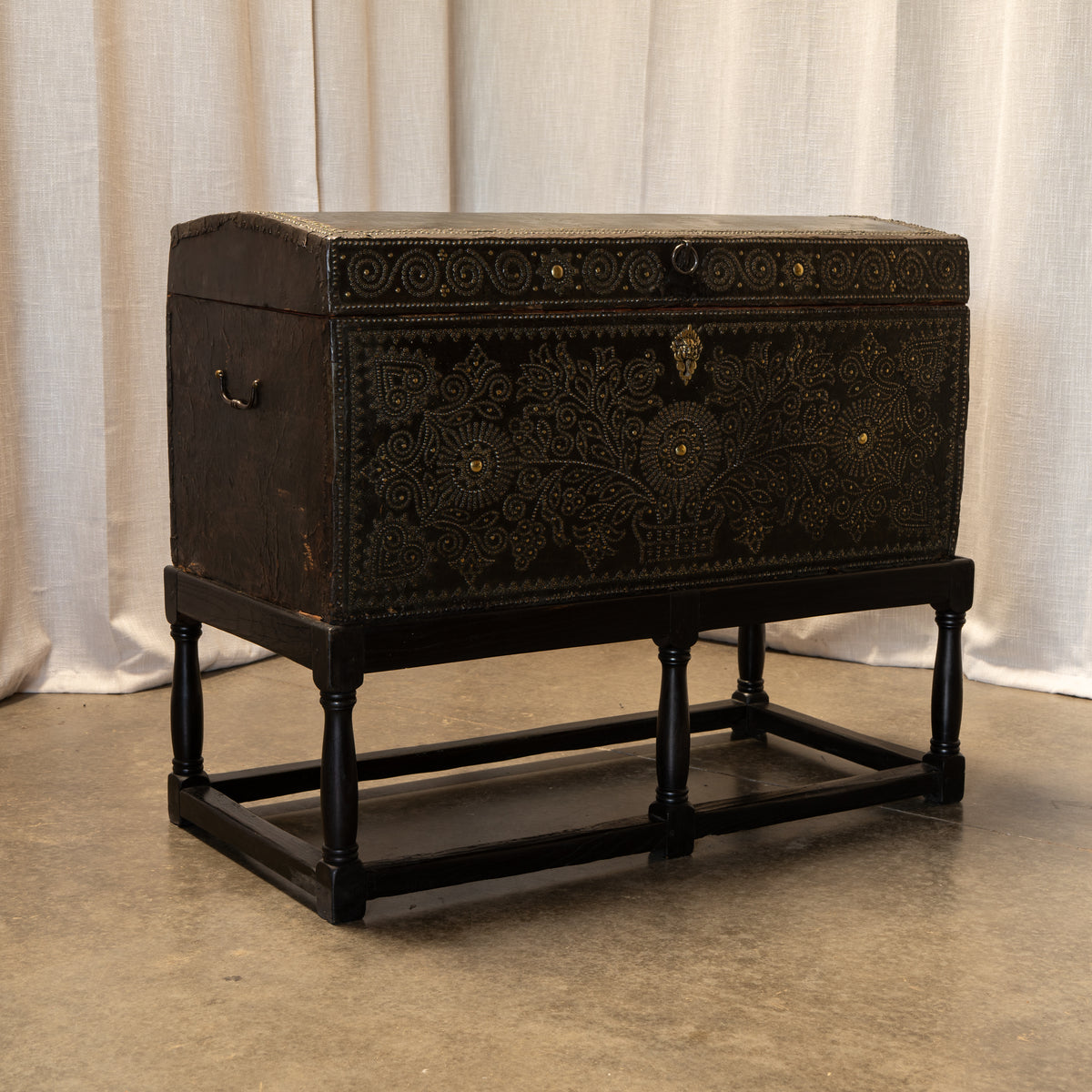 An antique dark brown wooden chest with intricate gold embossed floral patterns and a black wooden stand, set against a neutral draped fabric backdrop.
