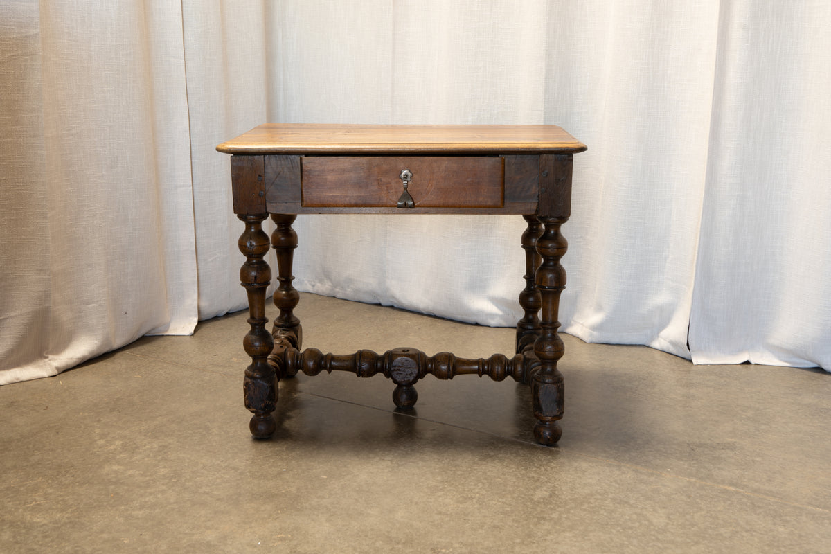 A rustic dark wood end table with a light natural wood top, a single drawer, and turned spindle-style legs, set against a draped white fabric backdrop.