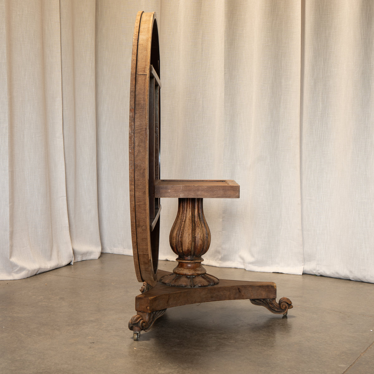 An antique wooden writing desk with a large circular back panel, turned pedestal base, and carved legs on casters, set against a draped cream curtain backdrop.