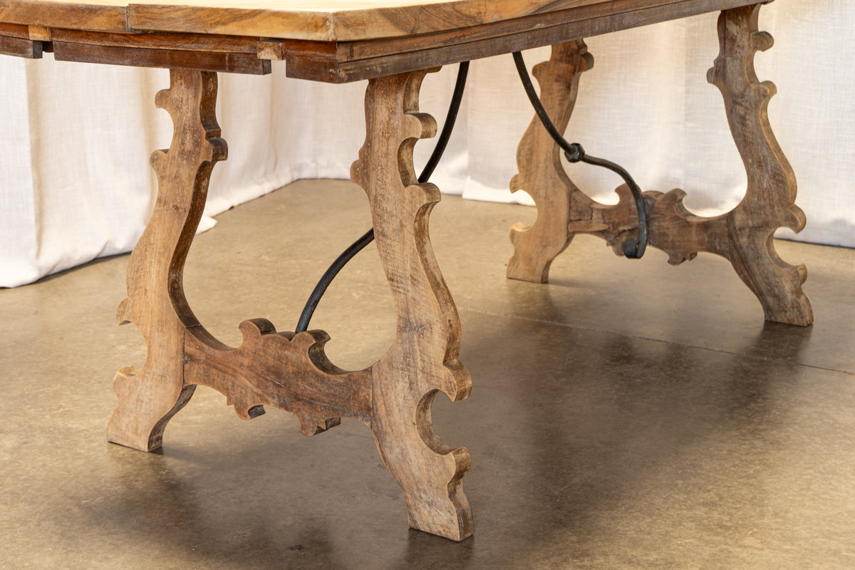 Close-up of an antique wooden table with ornate carved legs and a dark metal support cable, set against a neutral studio backdrop.