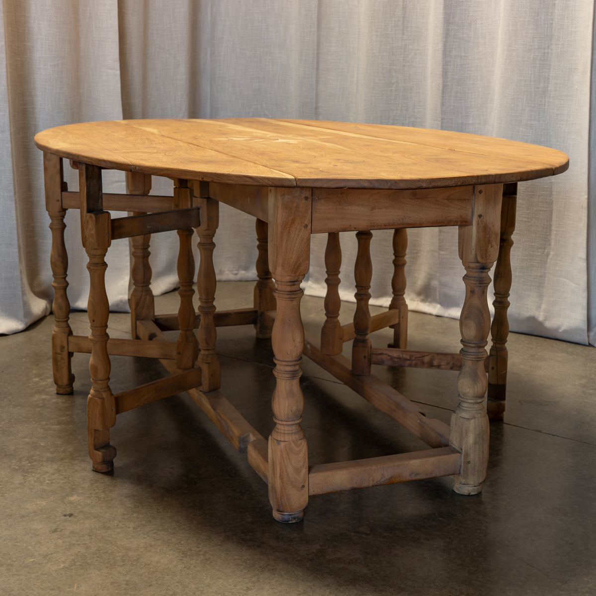 An antique wooden dining table with turned spindle legs and a drop-leaf design, photographed in front of a neutral curtain backdrop.