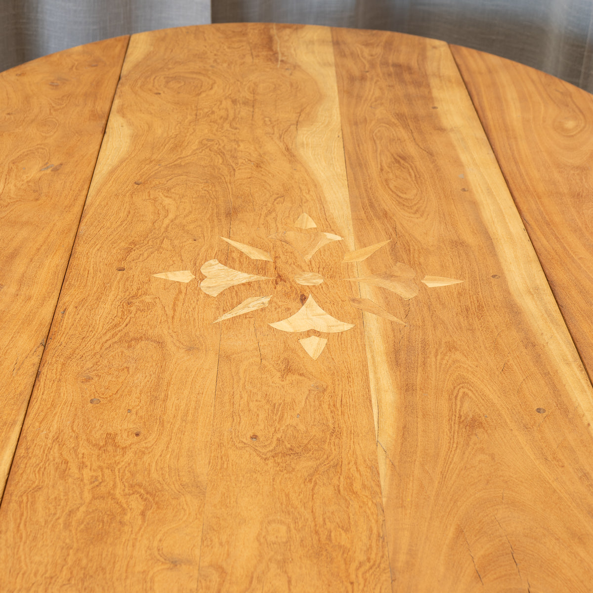 Close-up of a round wooden table top made of warm honey-toned wood planks with a light geometric inlay pattern in the center.
