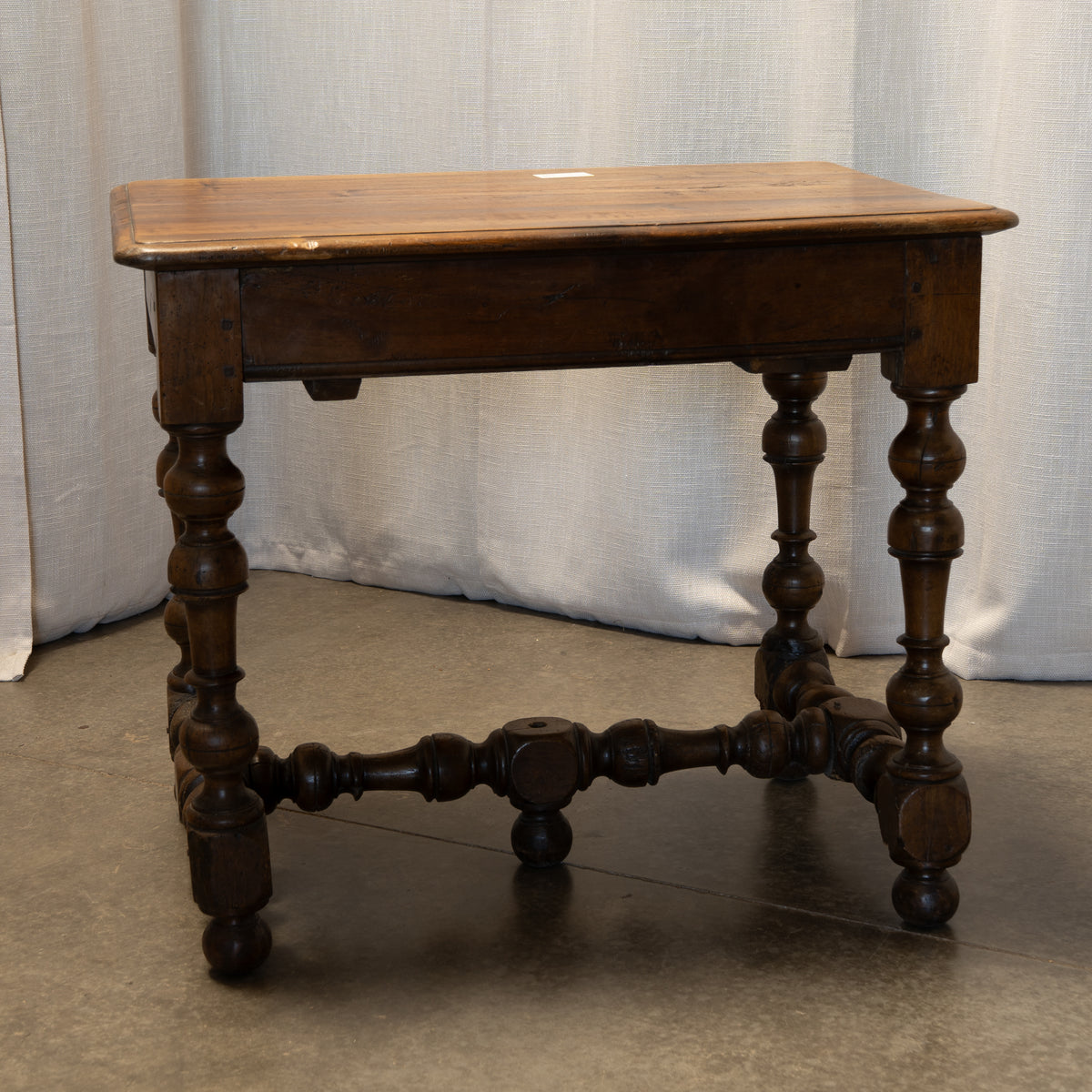 An antique dark brown wooden writing desk with turned ball-and-spindle legs and a central drawer, photographed against a light gray curtain backdrop.